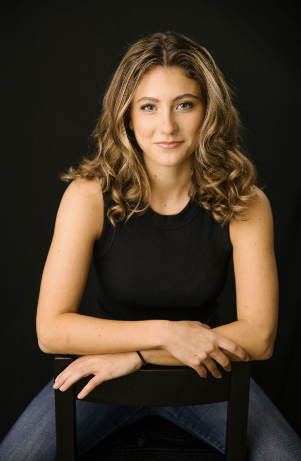 Tulia Henderson, theatrical headshot: seated, a slight smile, against a dark background.