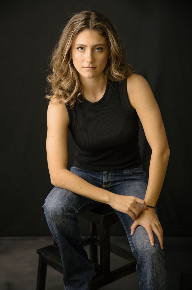 Tulia Henderson, dramatic headshot: seated on a stool, still and unsmiling, against a dark background.